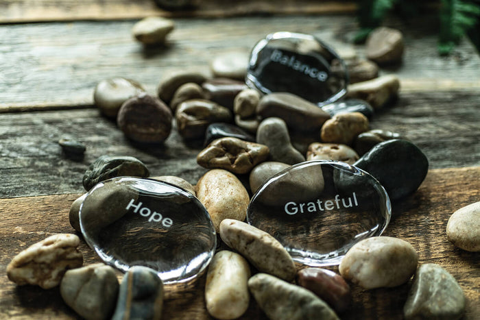 Black polished stone engraved with "Grateful" surrounded by natural pebbles on wooden surface.
