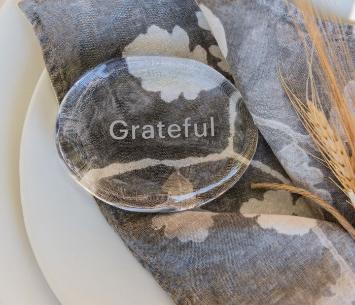 Clear glass stone engraved with "Grateful" resting on gray and white patterned fabric with dried wheat.