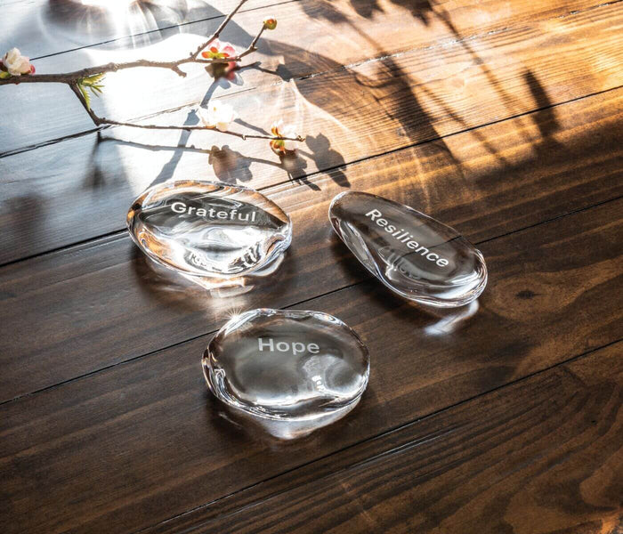 Three polished clear stones engraved with "Grateful," "Resilience," and "Hope" arranged on dark wood with dried flowers.