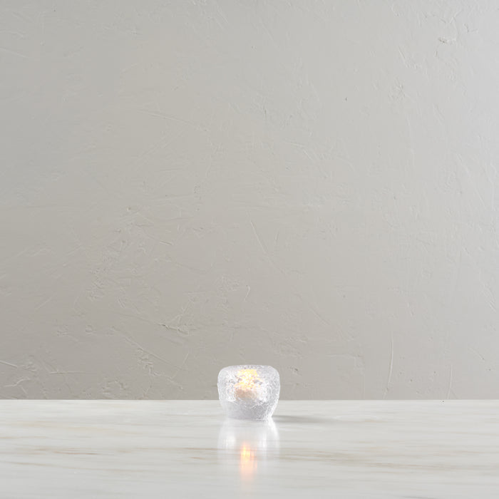 White spherical tealight holder with lit candle inside, displayed on light wooden surface.