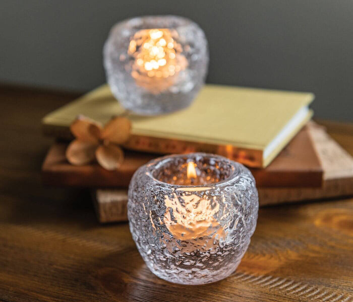Two clear textured glass tealight holders with lit candles, displayed on a wooden surface with books and dried flowers.