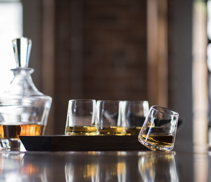 Four clear glass whiskey glasses with amber liquid on a dark wood base, with a matching decanter in background.