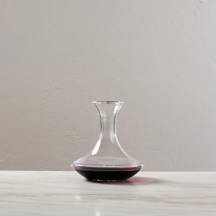 Clear glass wine decanter with wide base, narrow neck, and red wine inside, displayed on white surface.