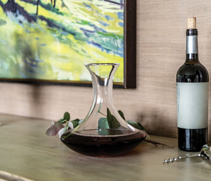 Clear glass wine decanter with wide base and tapered neck, displayed next to a wine bottle with greenery.