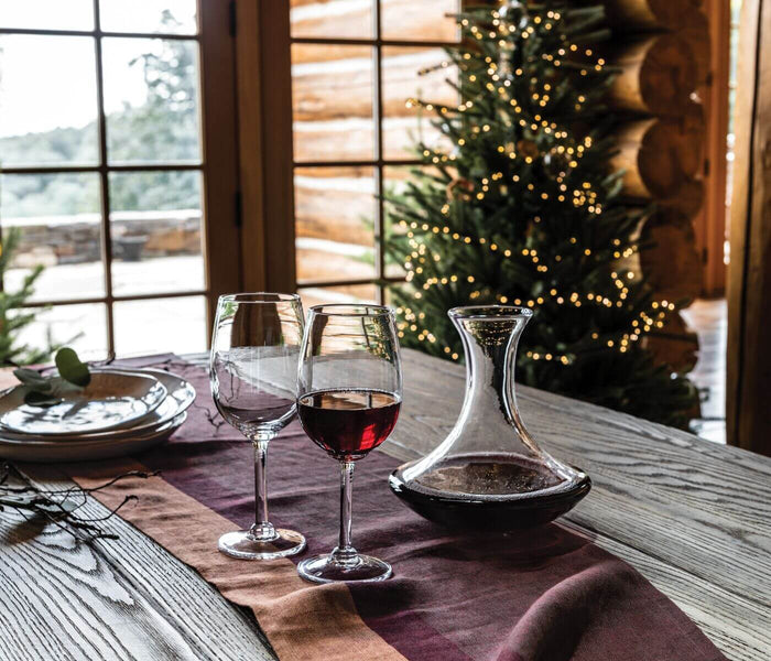 Clear glass decanter with curved spout next to two filled wine glasses on a dining table.