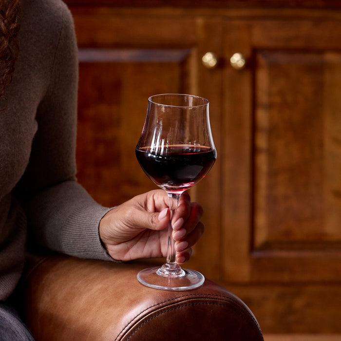 Wine glass with red wine held in hand, showing clear bowl on stemmed base with wooden interior background.