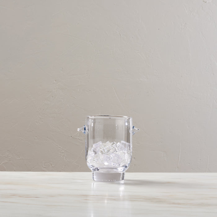 Clear glass ice bucket with two handles, filled with ice cubes.