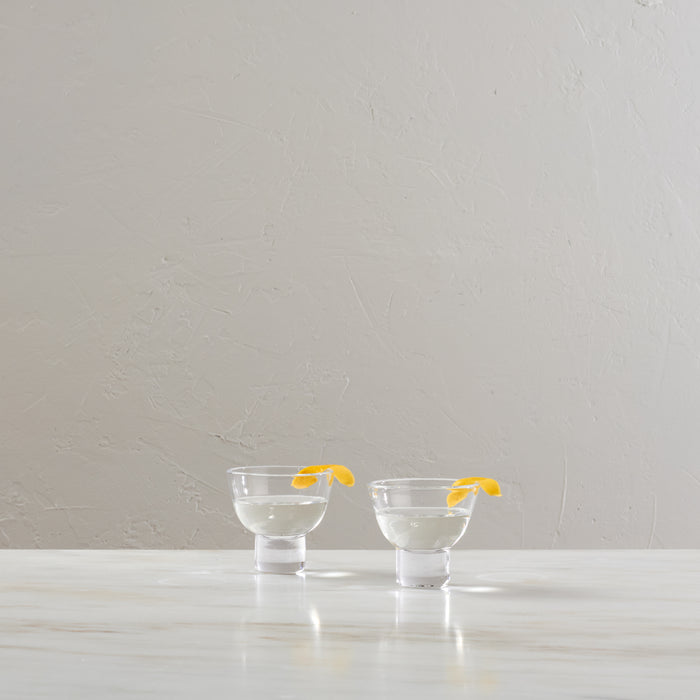 Two clear martini glasses filled with clear liquid and yellow lemon twists, displayed on a white surface.