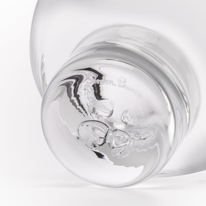 A close-up studio shot of a clear glass object, showing a rounded transparent body and a reflective base with the faint embossed brand name 'Simon Pearce'.