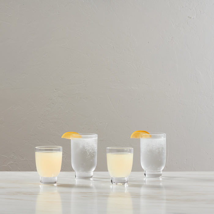 Four clear glass tumblers with pale yellow and sparkling beverages, two garnished with orange slices.