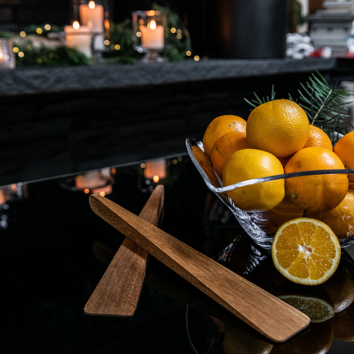 Two wooden salad servers with flat handles displayed next to a bowl of oranges and fresh greenery.