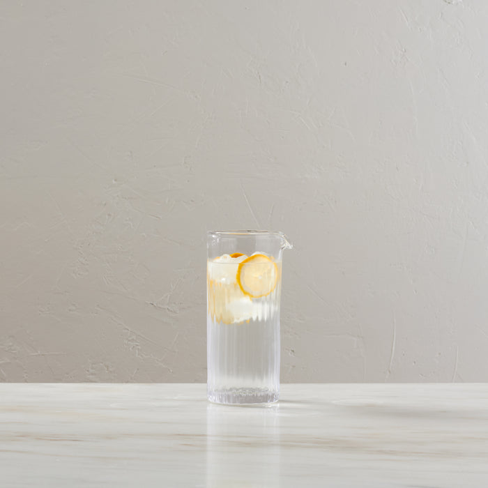 Tall clear glass pitcher with vertical ribbed texture, filled with water, ice, and lemon slices.