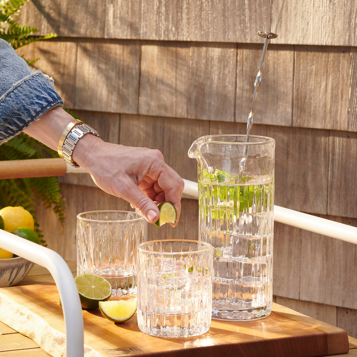 Clear glass pitcher with vertical ribbed texture being filled with liquid, displayed with matching drinking glasses on wooden