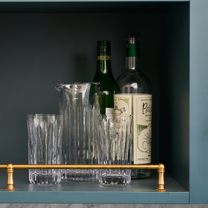 Two clear glass highball glasses with vertical ridge pattern, matching pitcher, and two bottles displayed on shelf.