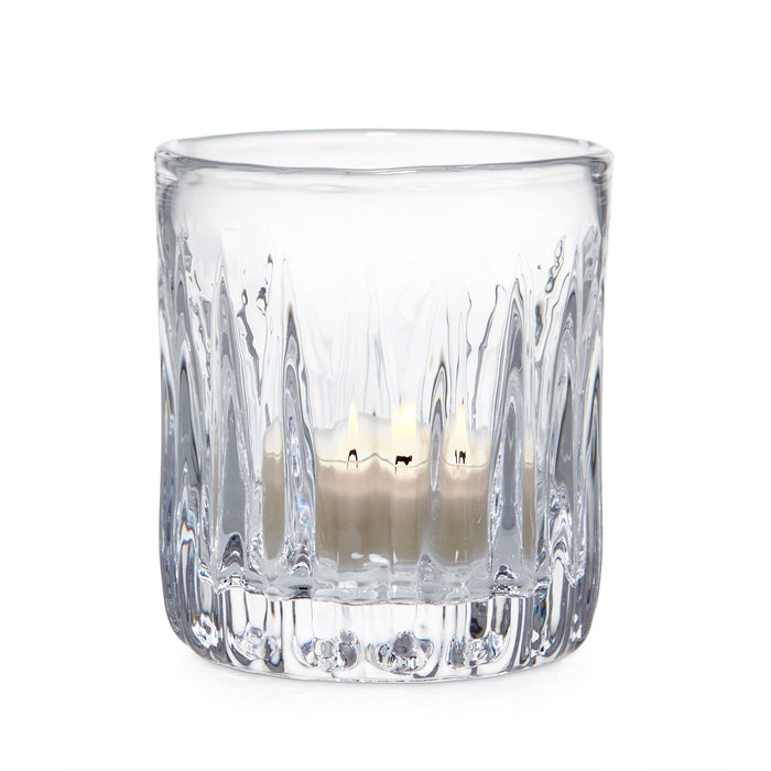 Clear glass tealight holder with vertical ribbed pattern containing lit cream-colored candle.