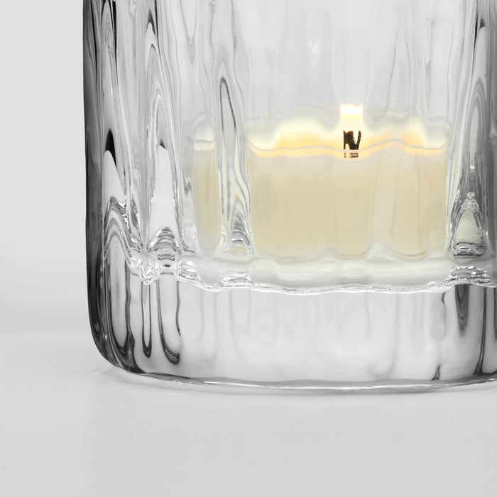 Clear glass container with textured ribbed sides holding a lit cream-colored tealight candle.