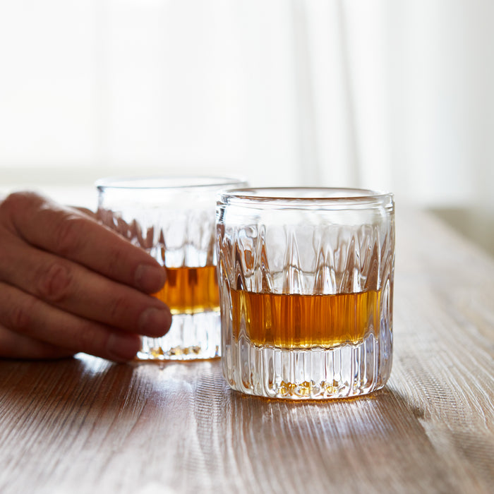 Two crystal rocks glasses filled with amber whiskey on a wooden surface with a hand reaching toward one glass.