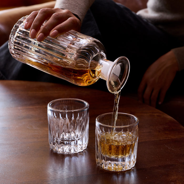 Amber whiskey being poured from a cut-glass bottle into two crystal glasses on a wooden surface.