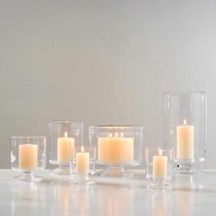 Set of seven clear glass hurricane candle holders in varying heights with lit cream-colored candles inside.