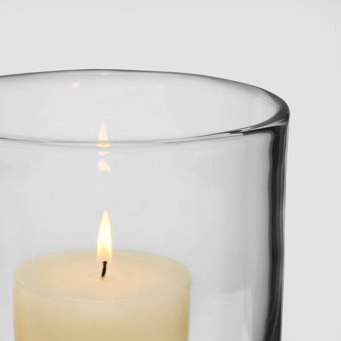 Cream-colored lit candle inside a clear glass hurricane with black rim.