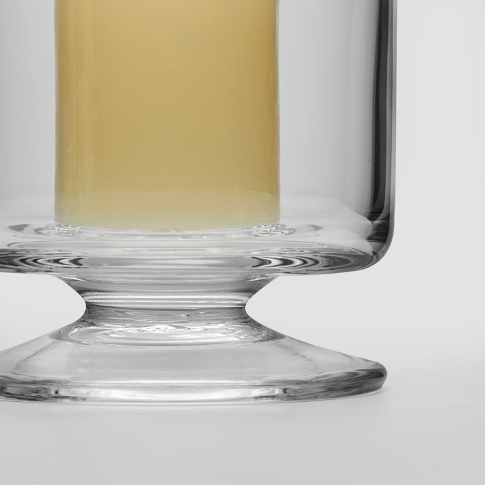 Clear glass hurricane candle holder with pedestal base and pale yellow liquid inside.