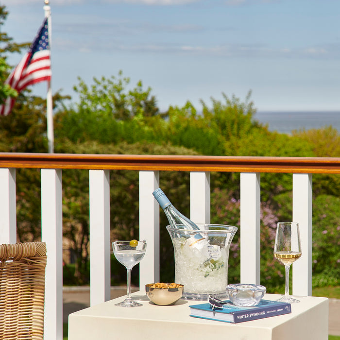 Clear glass ice bucket with metal handles containing ice and a chilled bottle on an outdoor table.