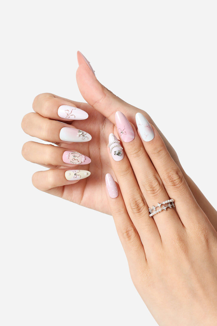 Almond press-on nails with pink and white ombre, featuring delicate bow designs, rhinestones, and pearl accents.