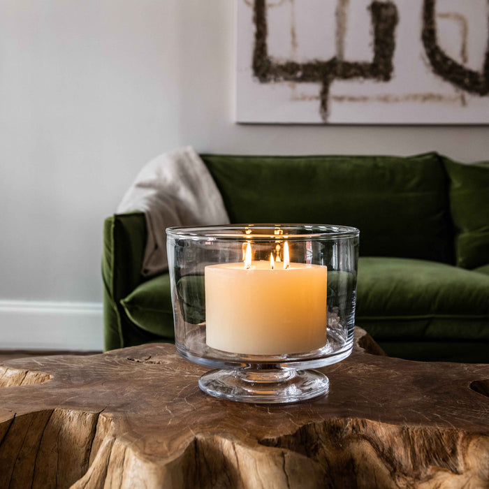 Ivory pillar candle with three lit wicks in a clear glass pedestal holder on a wooden table.