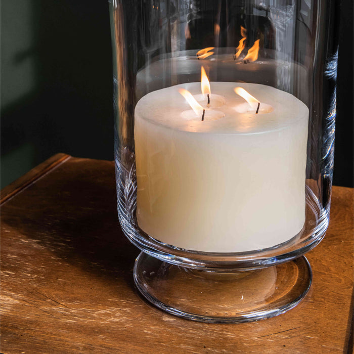 Ivory pillar candle with three lit wicks inside a clear glass hurricane holder on a wooden surface.