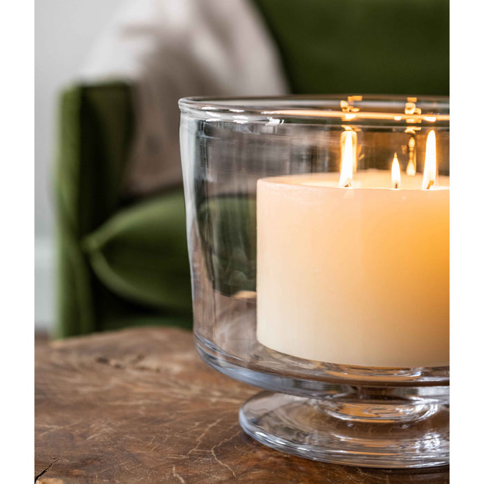Ivory pillar candle with three lit wicks in clear glass container on wooden surface.