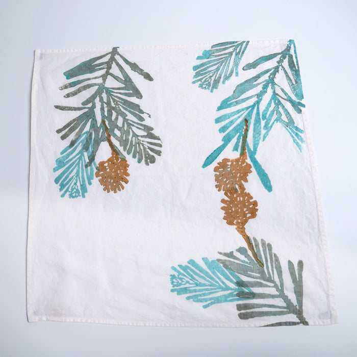 White linen napkin with pine branch and pinecone design in teal, gray, and gold.