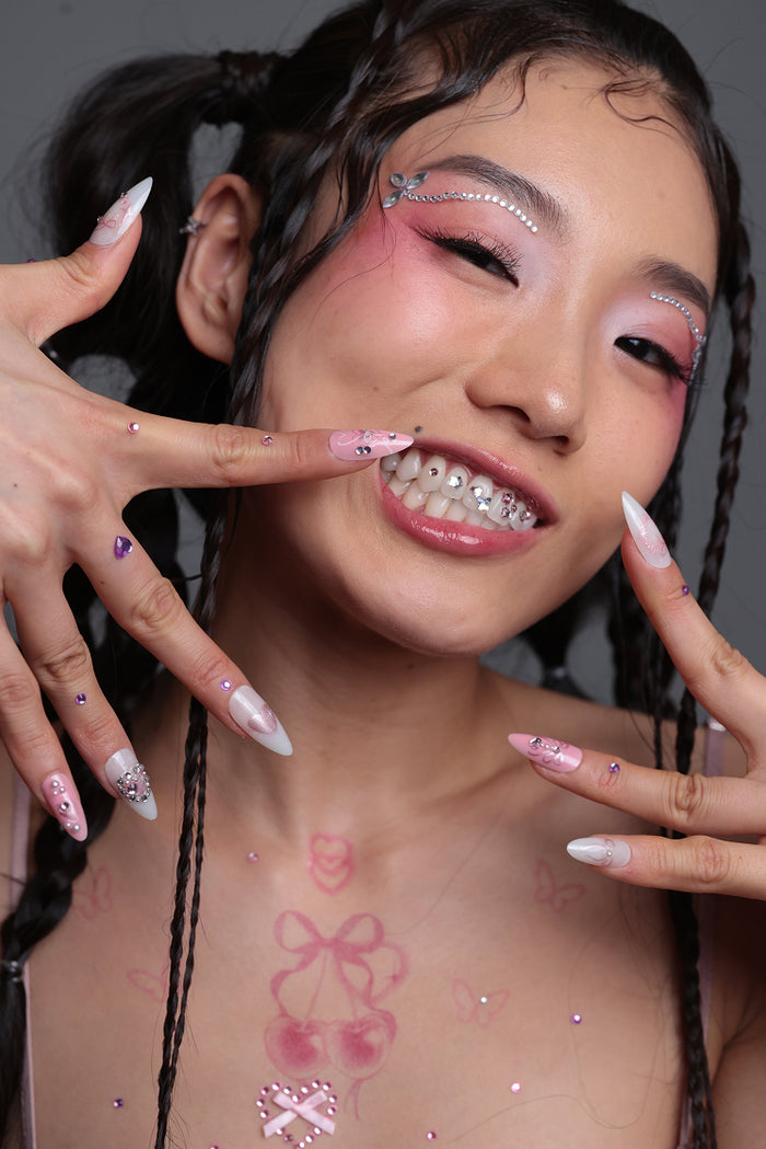 Young woman with pink eye makeup and facial gems smiling to show off sparkling tooth gems and decorated nails.