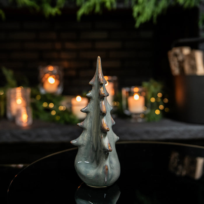 Gray ceramic evergreen tree figurine with pointed branches and rounded base, displayed with candles and greenery in backgroun