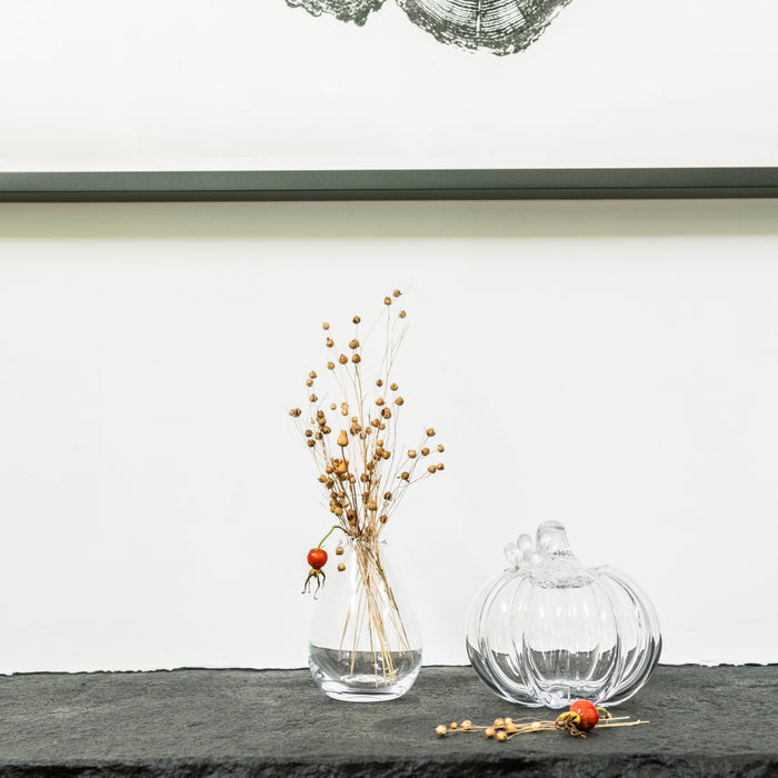 Clear glass vase with dried golden berry stems and single red berry, alongside pumpkin-shaped glass ornament on dark shelf.