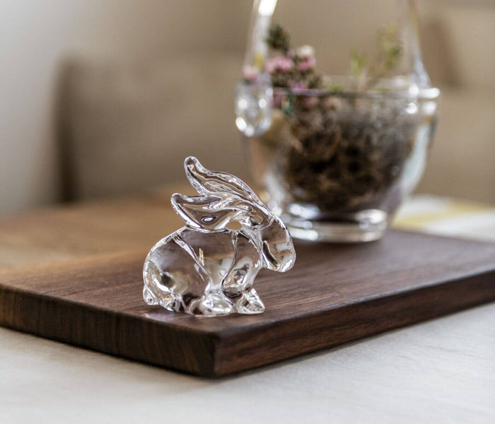 Clear glass rabbit figurine on wooden board with dried flowers in background.