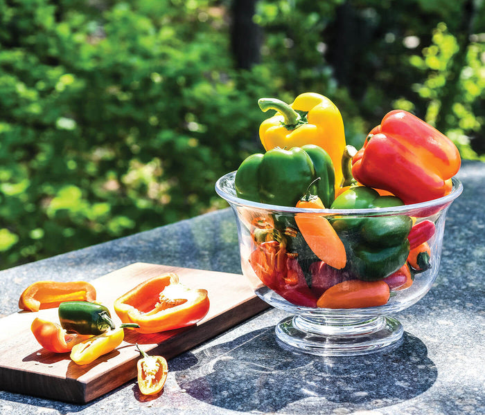 Clear glass pedestal bowl filled with red, yellow, and green bell peppers outdoors.