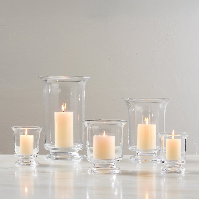 Five clear glass hurricane candle holders in varying heights containing lit cream-colored candles.