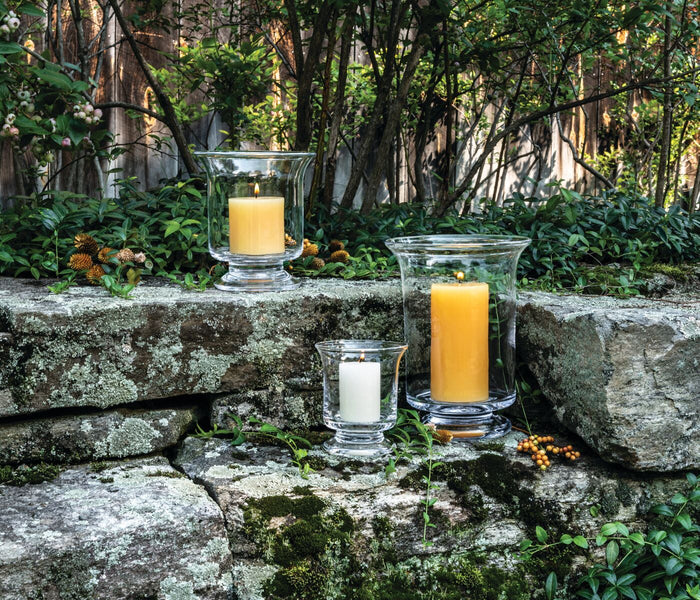 Three glass hurricane candles in varying heights with white and yellow wax, displayed on moss-covered stone outdoors.