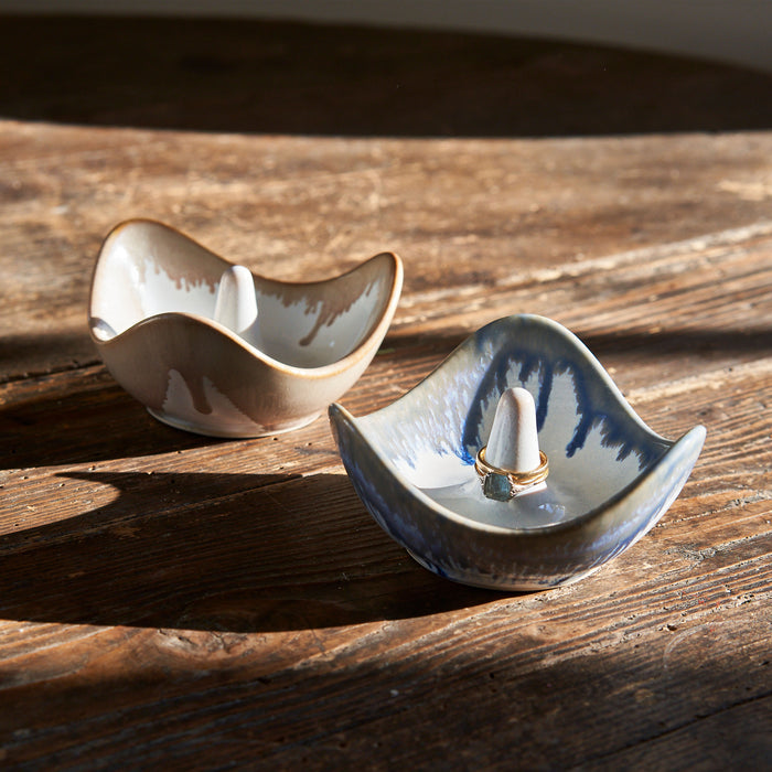 Two ceramic ring holders with curved wave shapes, one cream-colored and one blue-and-white, displayed on wooden surface.