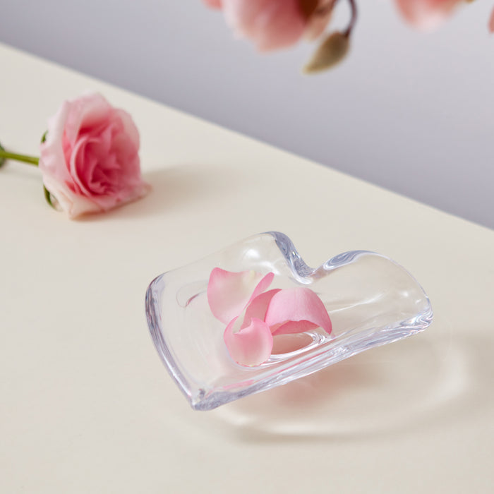 Clear heart-shaped dish with pink rose petals suspended inside, displayed on cream surface with fresh pink rose.
