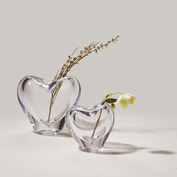 Two clear glass heart-shaped vases holding dried botanicals and fresh greenery.