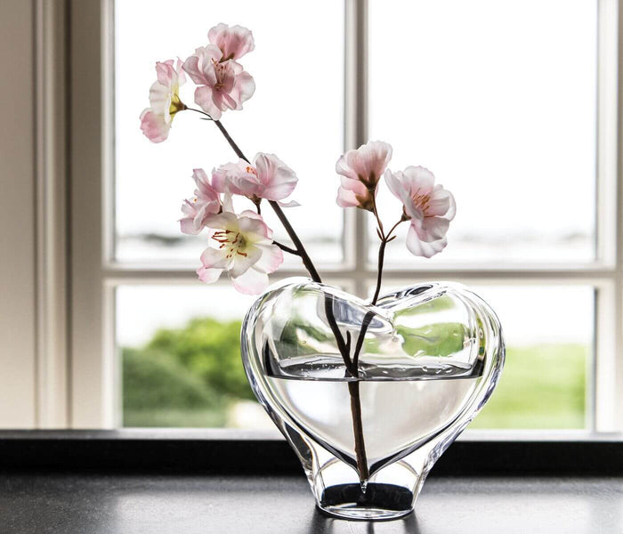 Clear glass heart-shaped vase with pink carnation branches and water inside, displayed on windowsill.