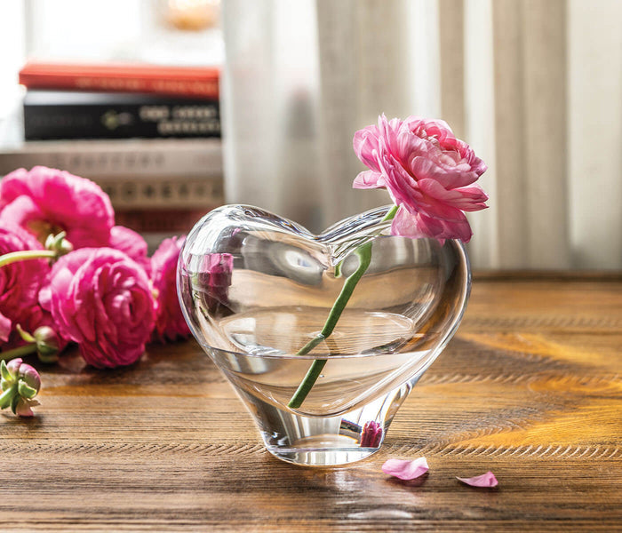 Clear glass heart-shaped vase with pink flower stem and water, displayed on wooden surface with books.