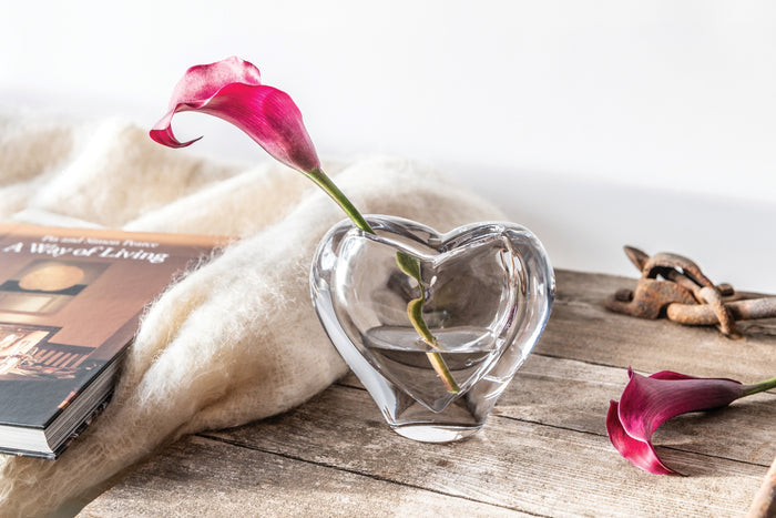 Heart-shaped clear glass vase with pink calla lily and green stem displayed on wooden surface.