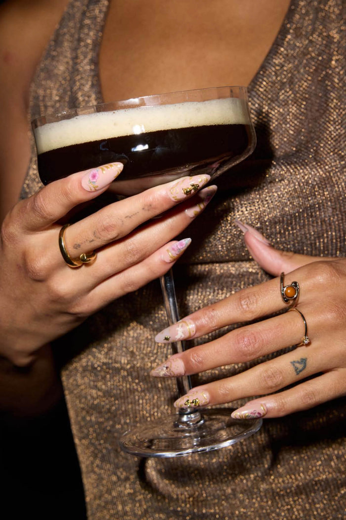 Rose gold rococo almond press-on nails with floral accents, holding a cocktail.
