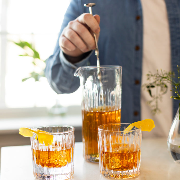 Stainless steel stirring spoon with long handle being used to stir amber liquid in a glass pitcher.