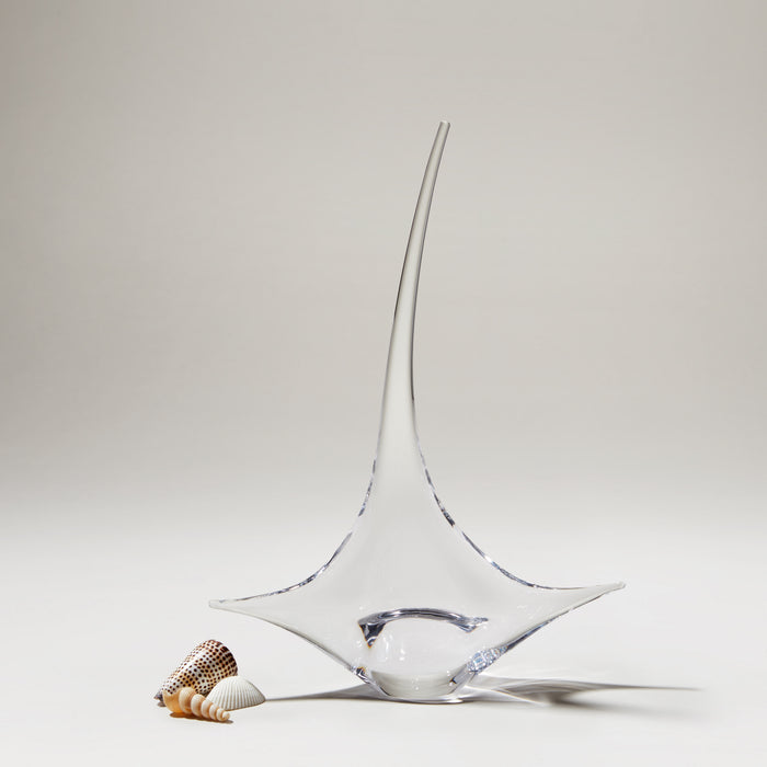 Clear glass sailboat sculpture with pointed mast and curved hull base, accompanied by small seashells.