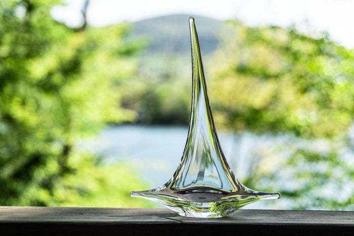 Clear glass sailboat figurine with pointed mast and wide base, displayed outdoors with blurred green foliage and water backgr