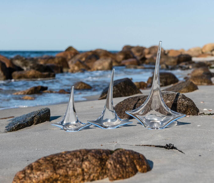 Three clear glass sailboat figurines on sandy beach with rocks and ocean in background.