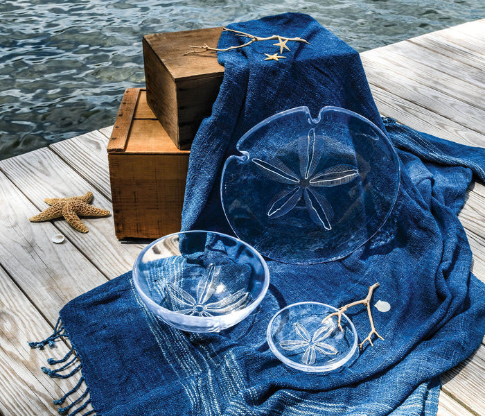 Blue glass bowls with sand dollar design, medium size, displayed on fabric near wooden box on dock.